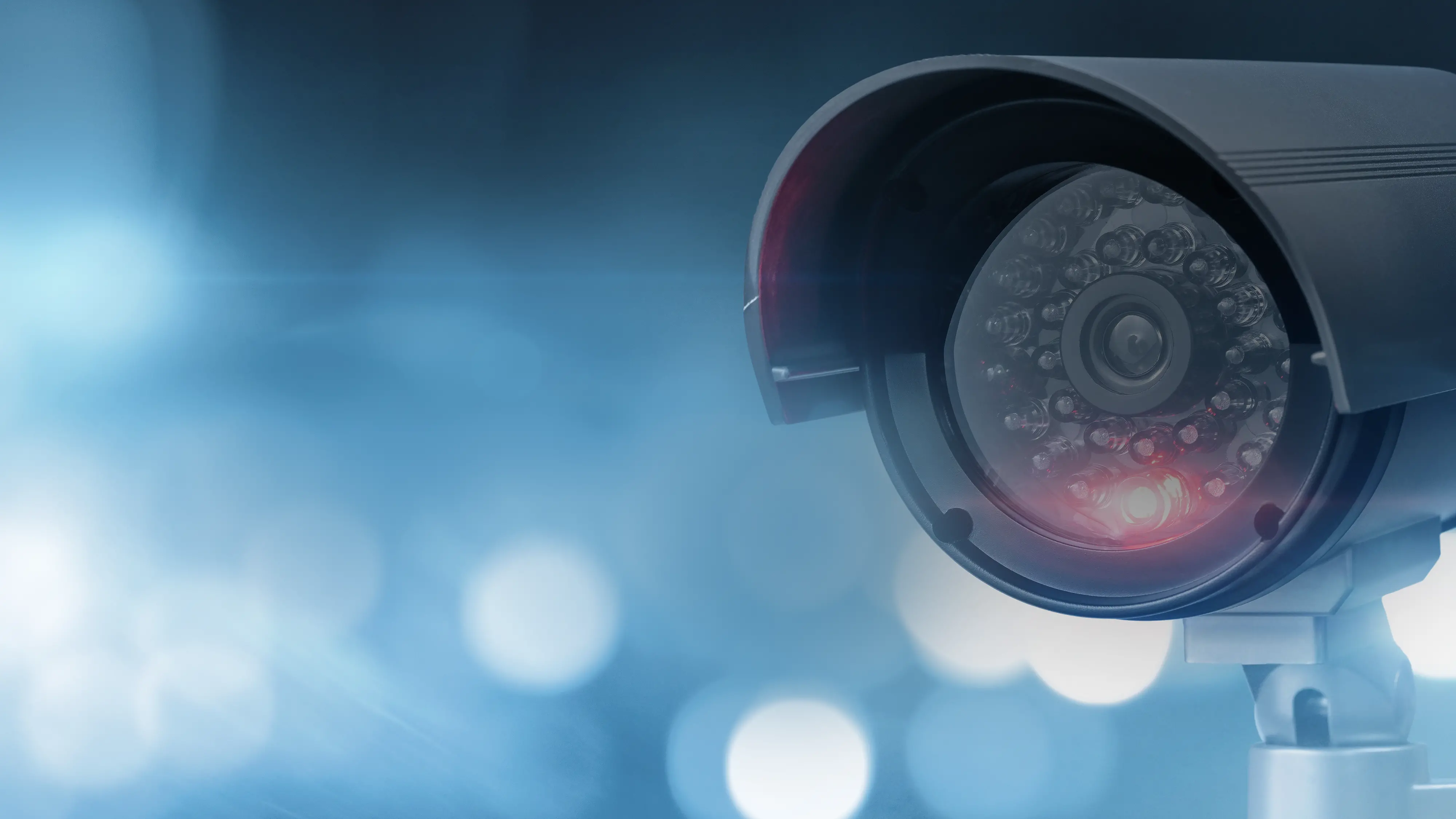 A black security camera with red lights is mounted on a ship. Blurred lights appear in the background.